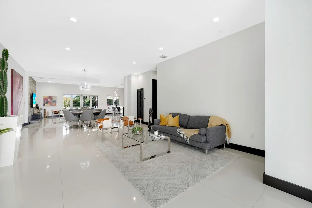 A stylish and welcoming interior view featuring a sofa with vibrant yellow cushions, a glass coffee table, large artwork on the wall, and a dining area in the background. The space feels lively and designed for social gatherings and family meals.