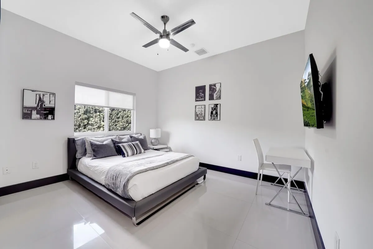 A contemporary and serene bedroom featuring a comfortable bed, decorative art above the headboard, a desk and chair ideal for remote work or study, and a wall-mounted TV. Natural light fills the room, enhancing its tranquility.