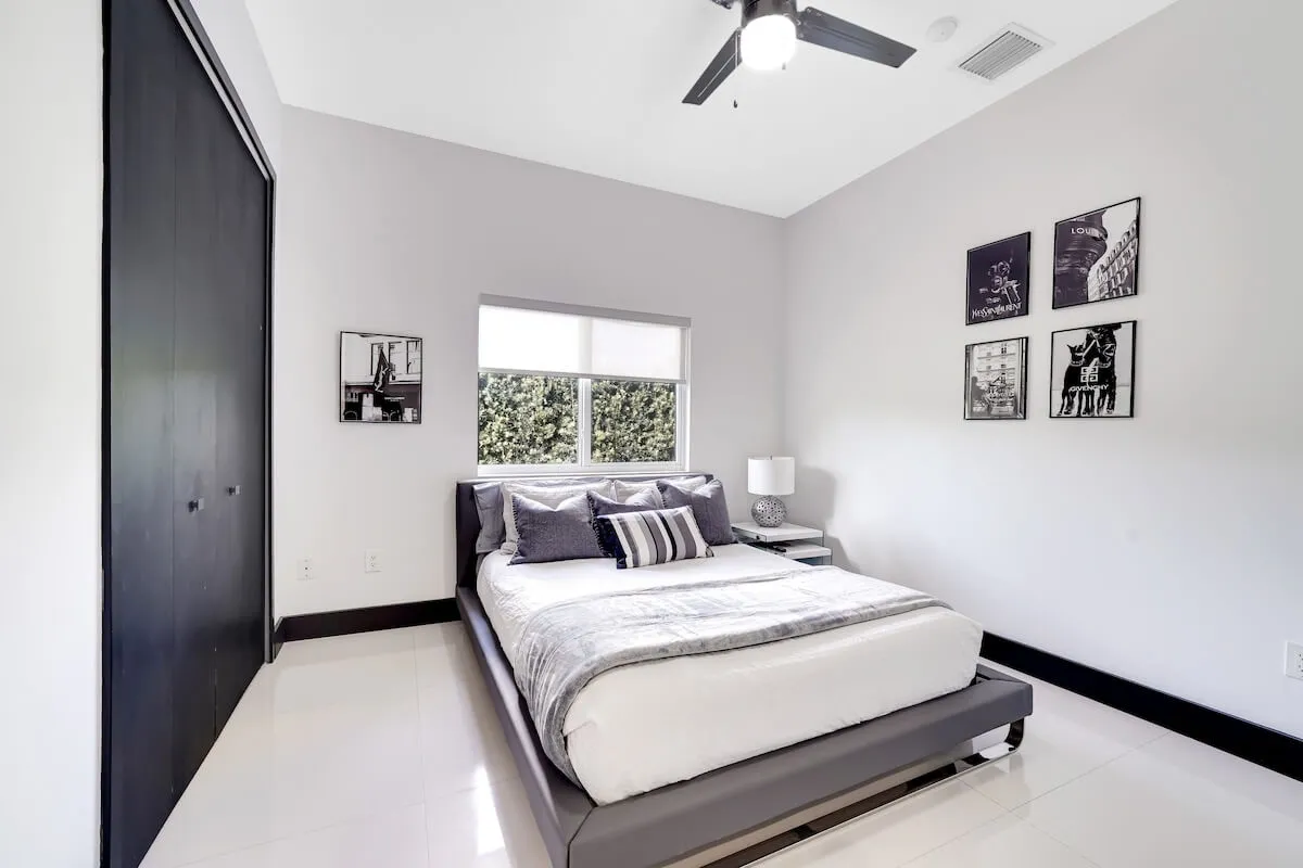 A minimalist bedroom with a bed dressed in grey and white bedding, a nightstand with lamp, black and white wall art, and a window allowing natural light to fill the space. The decor is calming and uncluttered, emphasizing restful comfort.