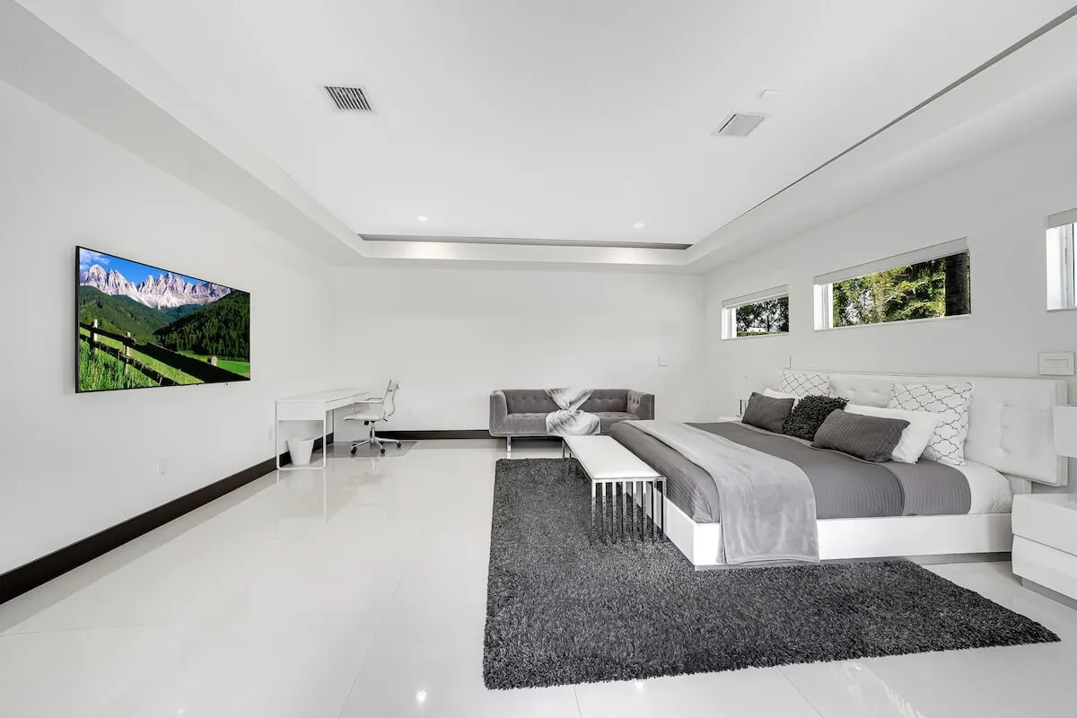 A serene and spacious bedroom featuring a king-sized bed with gray bedding, a modern gray sofa for lounging, a large TV mounted on the wall, and a white writing desk for work or study. The room is bright with natural light and offers a calming, uncluttered atmosphere.