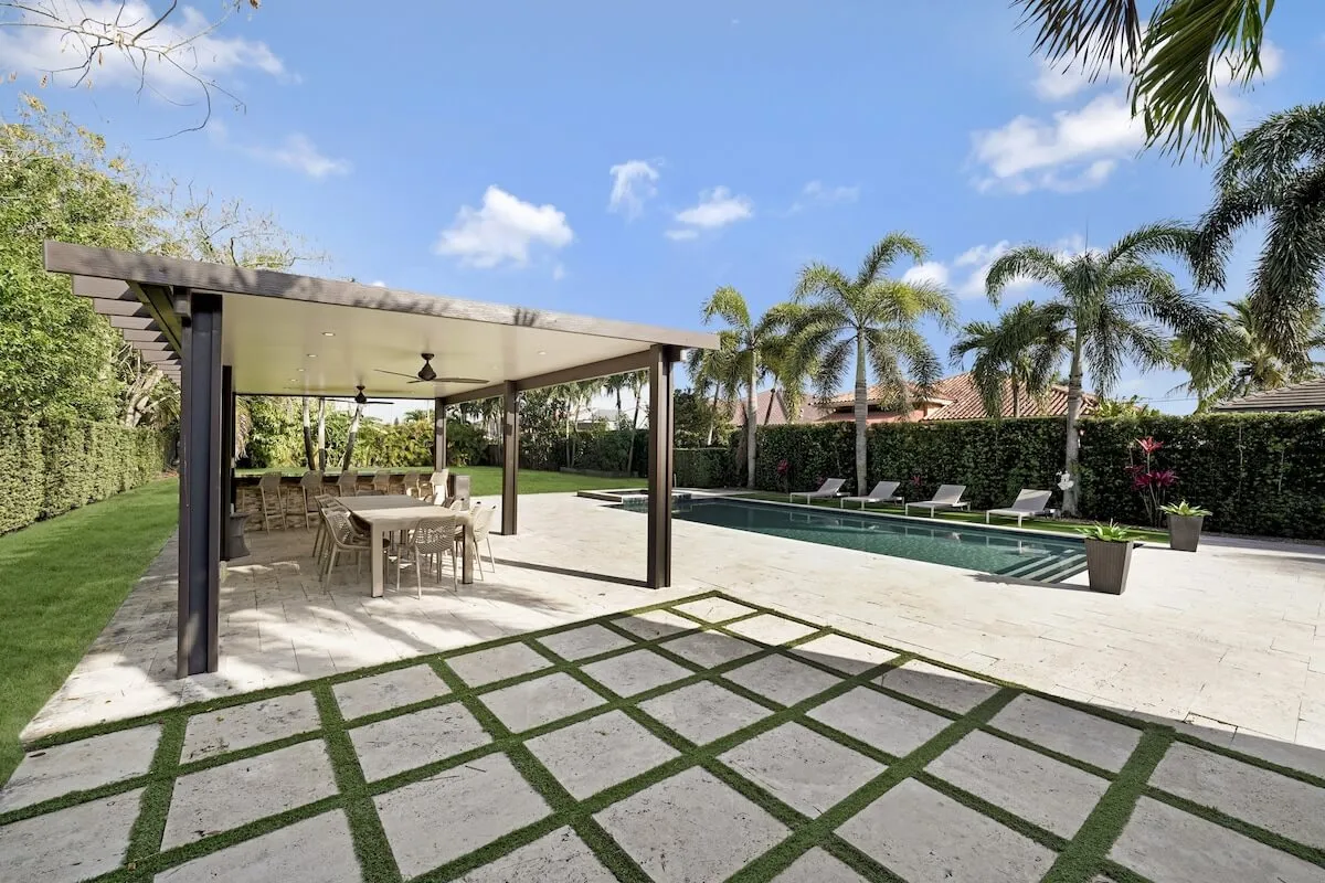 The image showcases a stunning Miami backyard oasis featuring a sparkling swimming pool surrounded by lush greenery, a sleek modern pergola shading an outdoor dining area, and ample space for relaxation or gatherings. The tranquil, resort-style setting is designed for both intimate evenings and lively family fun.