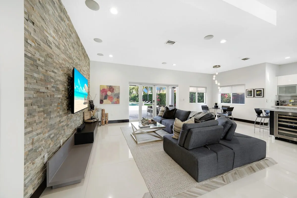 A spacious, contemporary interior featuring a large sectional sofa facing a television, a stone accent wall, and a dining area adjacent to glass doors that open to the outside. The space is bright and airy, with cohesive modern decor and plenty of room for family gatherings.