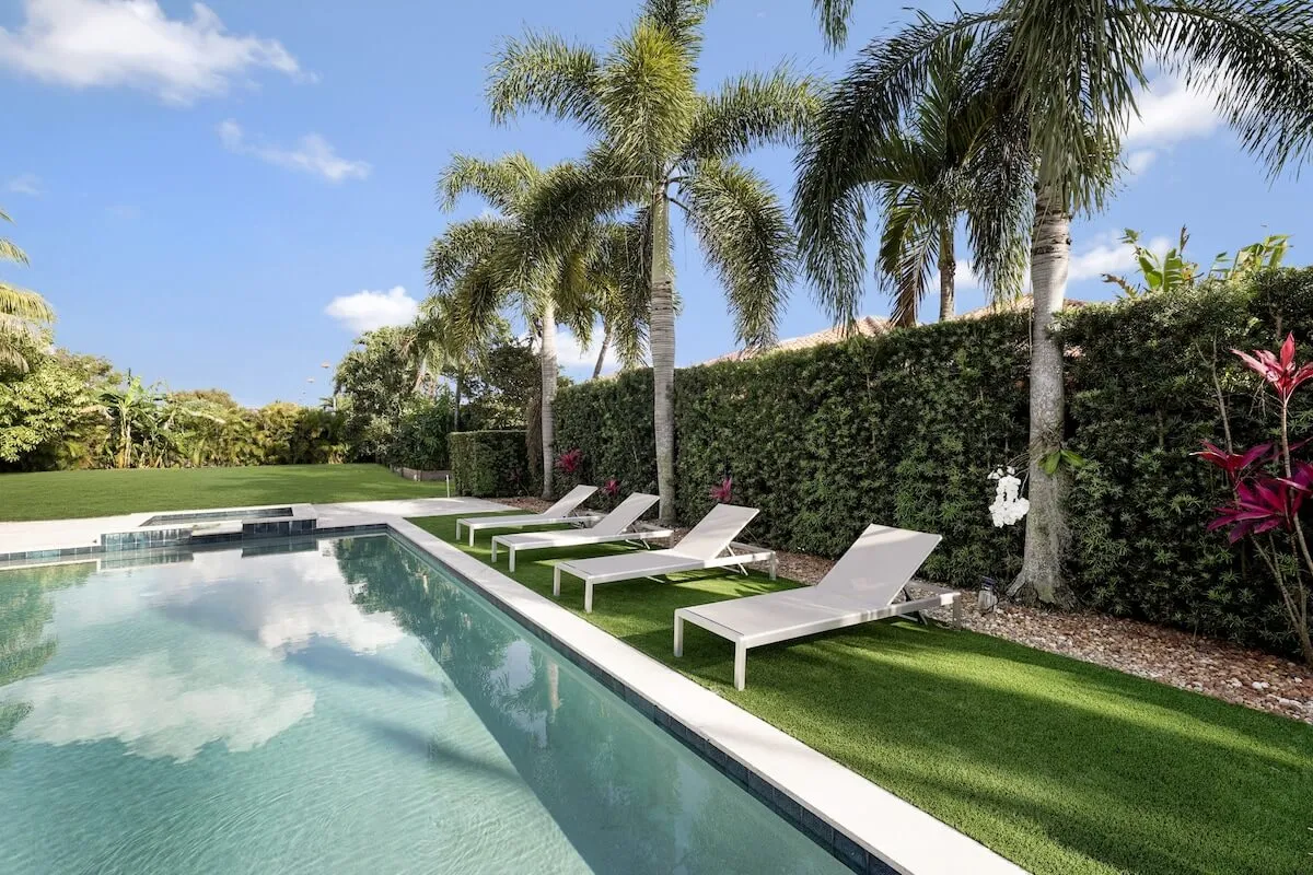 A beautifully landscaped backyard with a swimming pool, lounge chairs, palm trees, and a manicured garden. The area is set up for social gatherings and leisure time, offering ample space for sunbathing, swimming, or hosting a poolside event in a luxury setting.