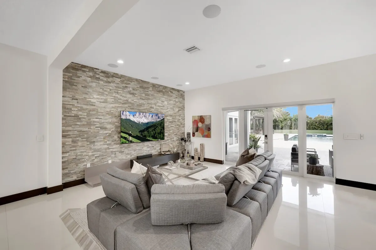 A bright, contemporary living room featuring a large sectional sofa, a stone feature wall with a mounted TV displaying a scenic mountain landscape, and glass sliding doors that let in natural light. The arrangement invites relaxation and group entertainment.