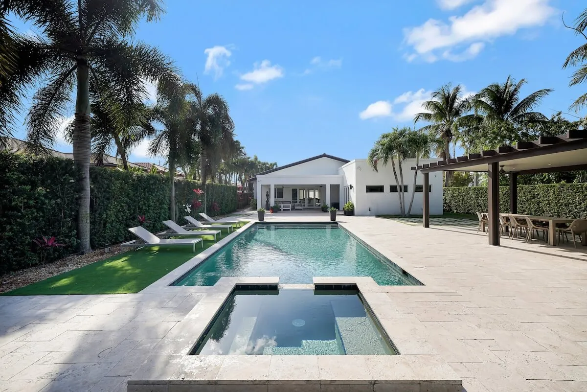 The image showcases a modern white Miami home with a sparkling rectangular pool and attached spa as its centerpiece. A row of lounge chairs sits beneath swaying palm trees, inviting guests to relax poolside. There's a stylish covered outdoor dining area perfect for group meals or entertaining, all set within a spacious, private backyard designed for both relaxation and small gatherings.