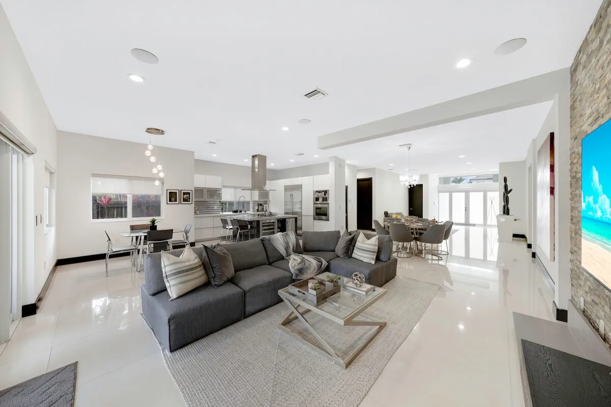 This image showcases a spacious open-plan living area featuring a large gray sectional sofa with decorative pillows, a modern kitchen island with bar stools and stainless steel appliances, a stone accent wall with a large flat-screen TV, and a dining area with a contemporary chandelier and round table. Sunlight fills the room through expansive windows, providing a bright and welcoming ambiance.