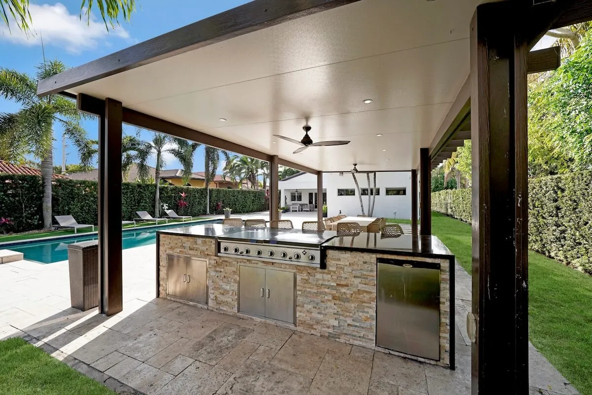 A well-equipped outdoor kitchen featuring a built-in grill and refrigerator sits under a covered patio with a ceiling fan. Beyond the kitchen, the pool and spa area blend seamlessly with the manicured backyard, which includes comfortable lounge chairs for relaxation.