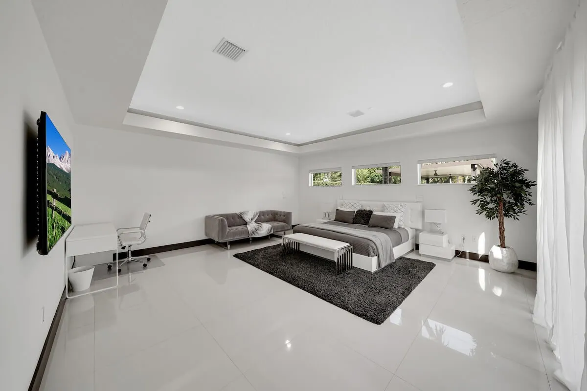 A serene master bedroom featuring a plush king-size bed with soft bedding, a large flat-screen TV mounted on the wall, a small modern sofa draped with a throw blanket, and a white desk with an ergonomic chair. The space is flooded with natural light, creating an inviting and tranquil atmosphere.