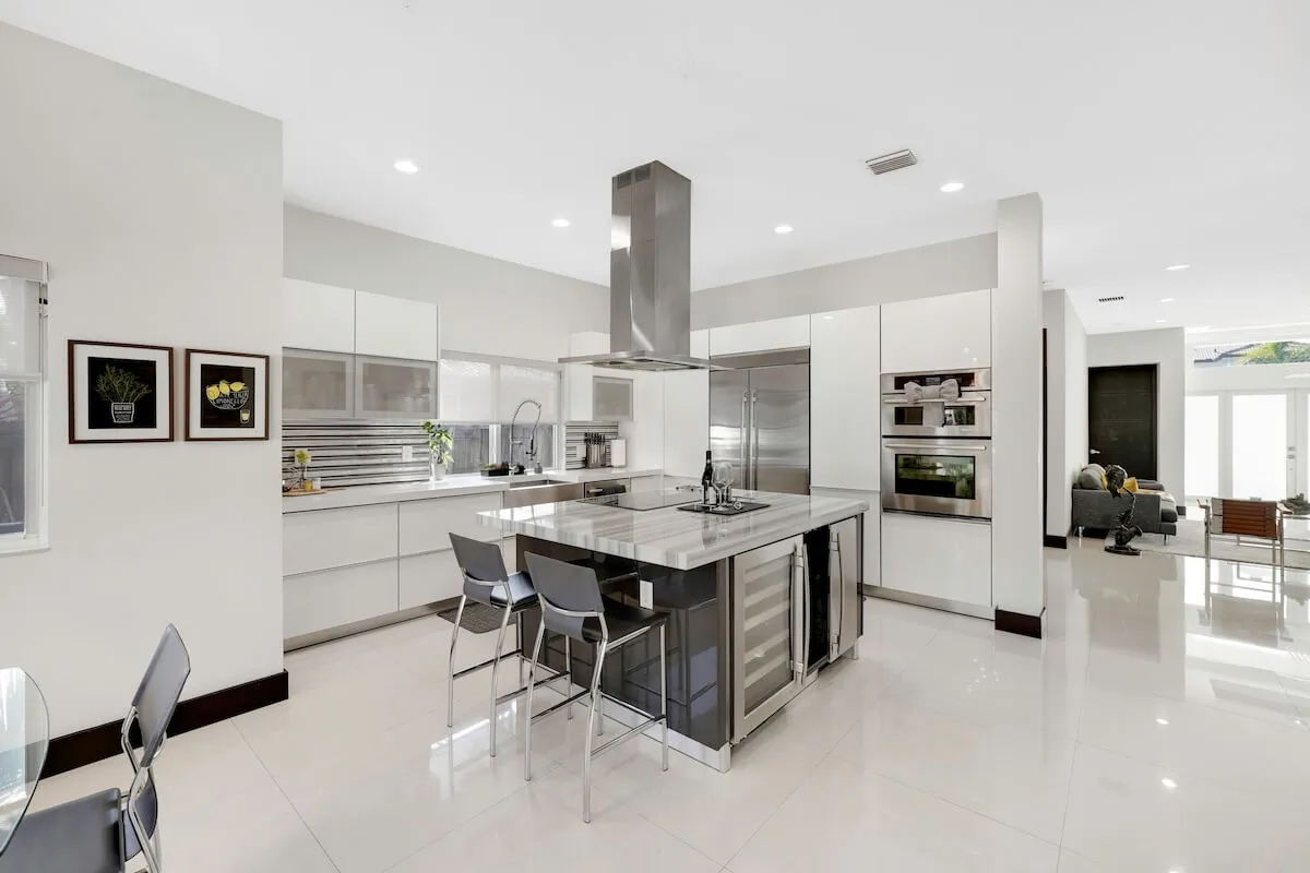 A bright, modern kitchen centered around a large island with built-in stovetop and bar seating. Stainless steel double-door refrigerator and double ovens line the wall, while expansive windows flood the space with natural light. The overall layout encourages both meal preparation and social interaction, with plenty of space for multiple people to gather.