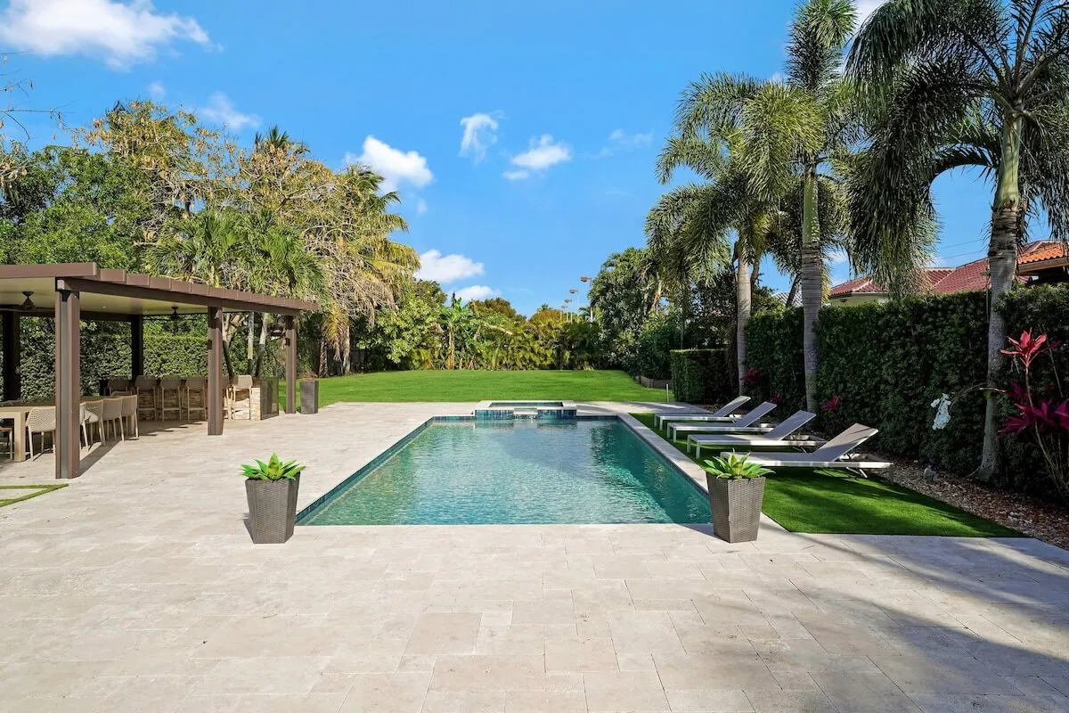 A vibrant exterior view of a backyard oasis featuring a rectangular pool with clear water, several poolside lounge chairs, a covered outdoor kitchen and dining area, and a large green backyard with lush landscaping. The scene radiates energy and sociability, with ample space for groups to gather, swim, cook, and dine outdoors.