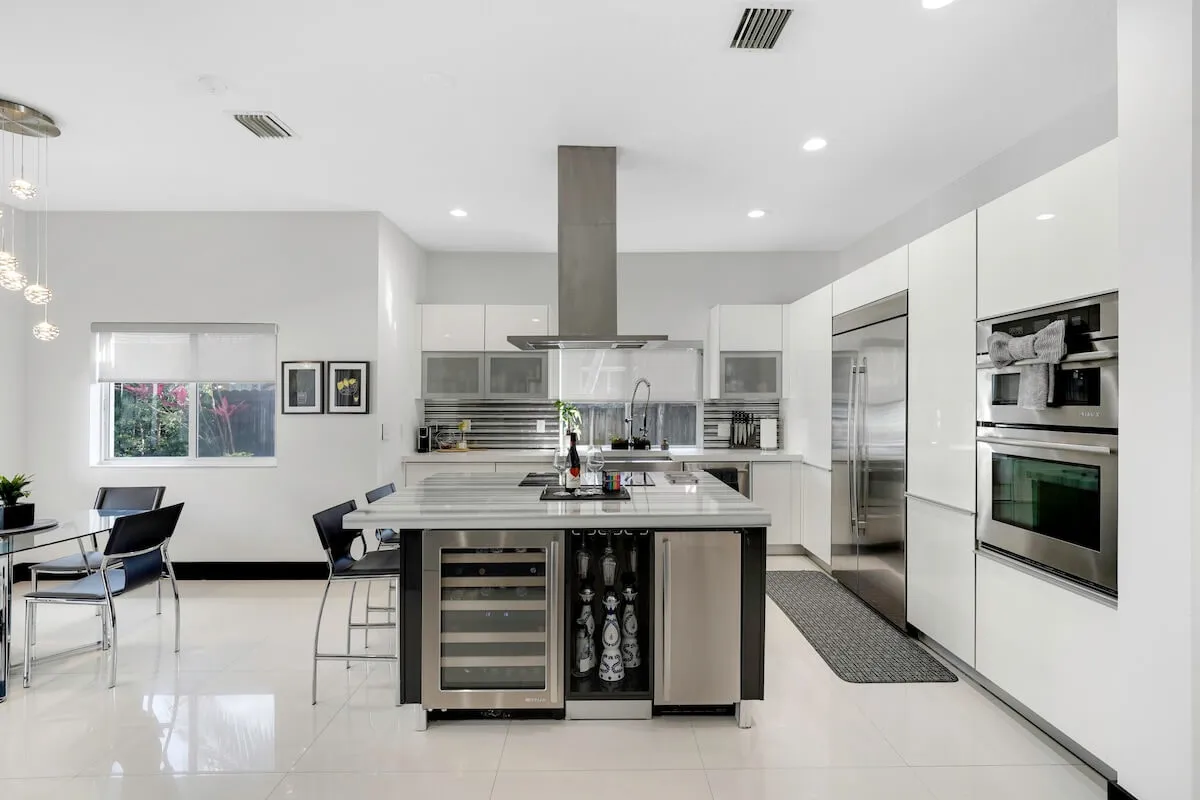 A stylish kitchen island with a built-in wine cooler and ample storage anchors this open-plan space. Stainless steel appliances, including double ovens and a refrigerator, are set against sleek white cabinetry. A dining area sits adjacent, perfect for sharing meals and stories.