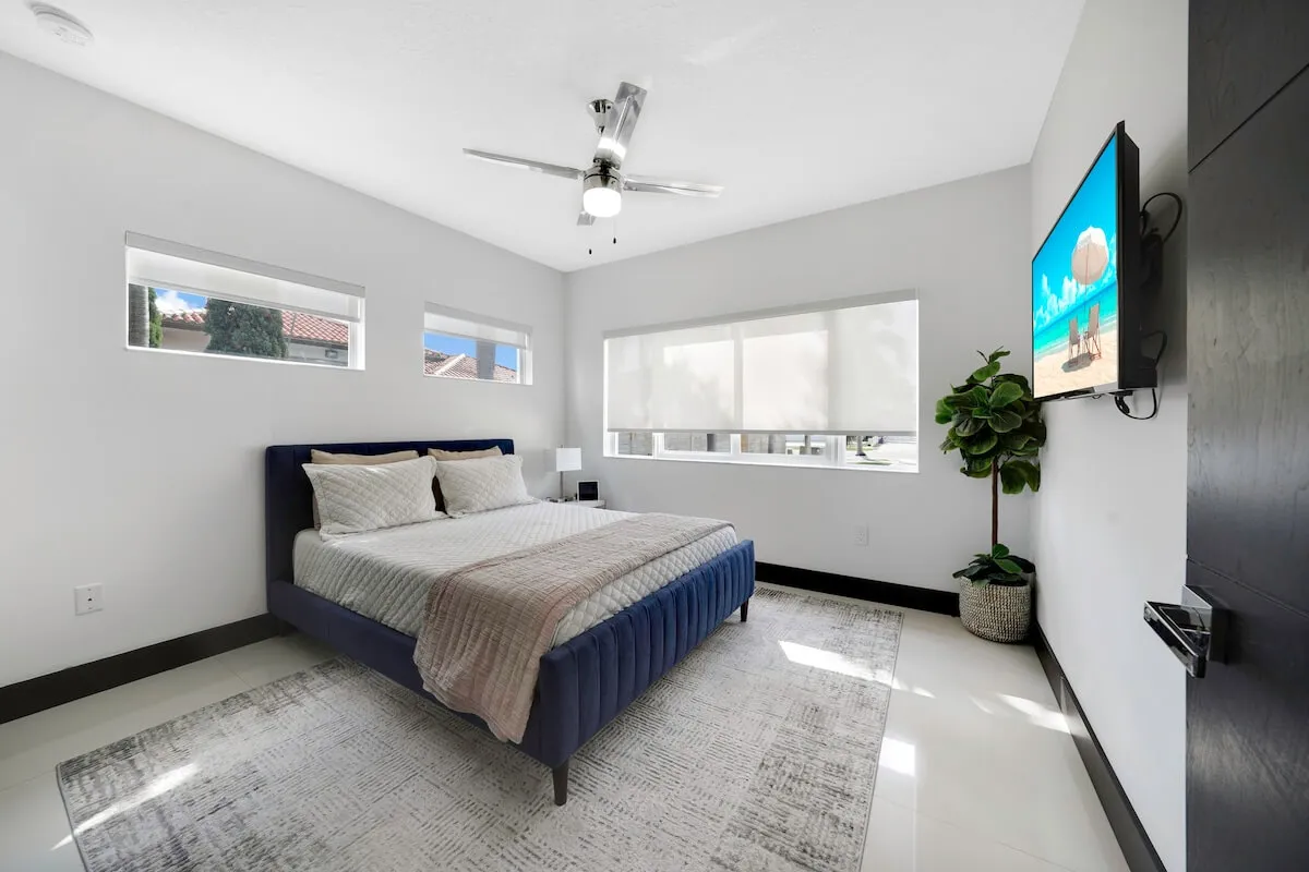 A stylish bedroom featuring a plush king-sized bed with a navy blue upholstered frame, large windows filtering in natural light, a wall-mounted flat-screen TV displaying a beach scene, and a touch of greenery from an indoor potted plant. The mood is restful and inviting.