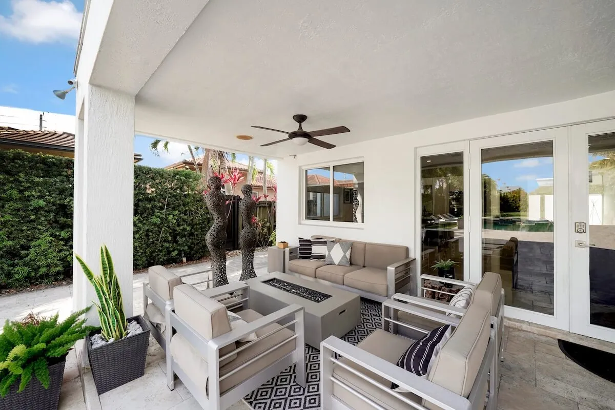 A cozy outdoor seating area on the patio, featuring cushioned chairs and a sofa arranged around a central fire pit table. Decorative metal sculptures and a lush green hedge provide privacy and visual interest, making the space ideal for intimate gatherings or relaxed socializing.