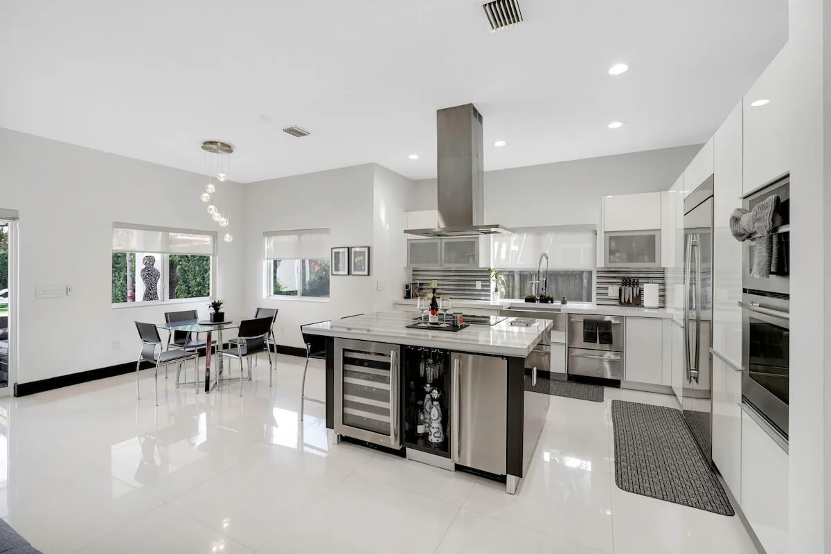 A contemporary kitchen features a large island with a built-in wine cooler and cooktop, stainless steel double oven and refrigerator, and an adjacent dining area with a modern glass table and chairs. Sleek, bright finishes and pendant lighting create an airy, sophisticated vibe ideal for family gatherings or entertaining guests.