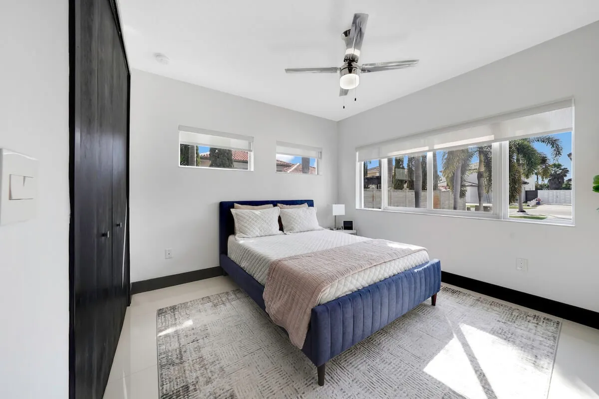 A bright and restful bedroom with a blue upholstered bed topped with white and beige linens, large sunlit windows, dark wood closet doors, and a ceiling fan with a gentle light. The room exudes calmness and privacy, perfect for kids or guests.