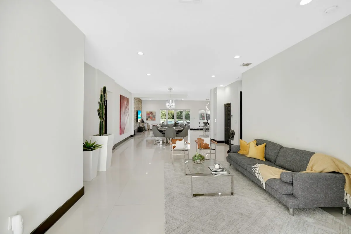 A calm and inviting living space featuring a gray sectional sofa with yellow and beige throw pillows and a blanket, a glass coffee table with decorative items, a dining area with contemporary chairs, and vibrant wall art and potted plants. The room exudes warmth and is well-lit by natural light.