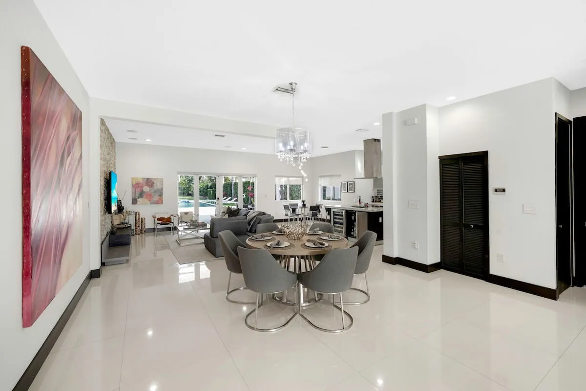 A bright, stylish interior with a round dining table and modern chairs under a contemporary chandelier, comfortable living room seating facing a TV, and large glass sliding doors that reveal the pool area outside. The space is open and airy, with natural light streaming in, emphasizing the connection between indoor and outdoor living.