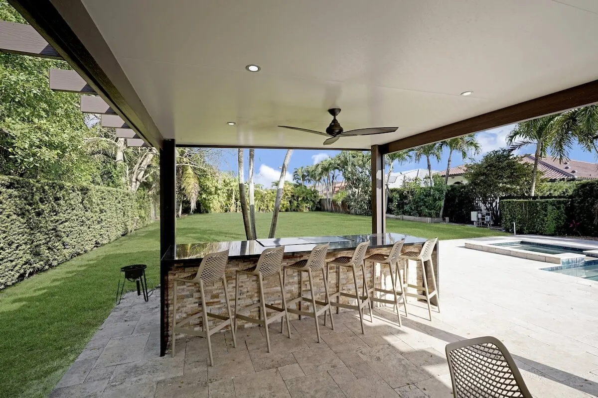 This wide-angle exterior photo shows a backyard bar with high chairs under a covered patio, a swimming pool with spa, and a large lawn area. The setting is designed for socializing, outdoor dining, and family celebrations, with palms and hedges providing privacy.
