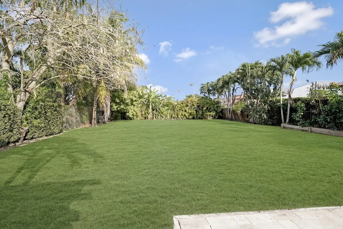 A large, open expanse of green grass is framed by lush tropical plants and palm trees under a clear blue sky. The space is ideal for children’s games, picnics, or laid-back family gatherings, with an inviting and serene atmosphere.