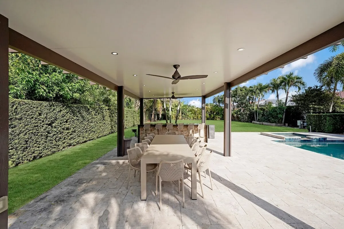 A long dining table with multiple chairs is set on a covered patio, complete with ceiling fans for comfort. The pool and spa are visible nearby, bordered by a well-manicured lawn and tall hedges for privacy.