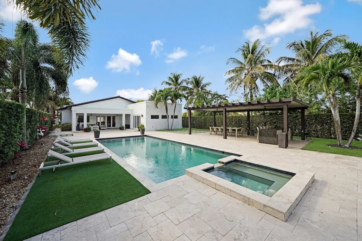 A wide-angle view of the backyard featuring a modern rectangular pool with an integrated spa, a covered outdoor kitchen and dining area, and a white contemporary house with large sliding doors. Palm trees and lush greenery surround the backyard, creating a tranquil, resort-like setting ideal for gatherings.