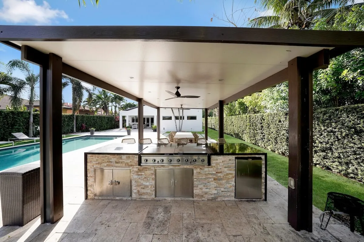 A wide-angle shot of a lush backyard showing a long swimming pool, a covered outdoor kitchen with a built-in grill, and a patio dining table set beneath the cover. Tall palm trees and hedges provide privacy and a tropical vibe, reinforcing a sense of leisure and exclusivity.