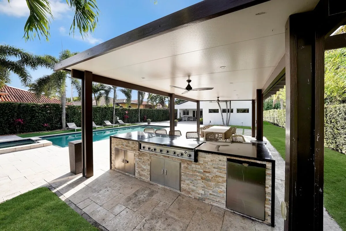 This vibrant exterior image features a well-equipped outdoor kitchen with a grill and countertop, a covered dining area with a table and chairs, a sparkling swimming pool lined with lounge chairs, and a lush, well-maintained lawn and hedges. The scene is lively and inviting, emphasizing family-friendly amenities and outdoor living spaces.
