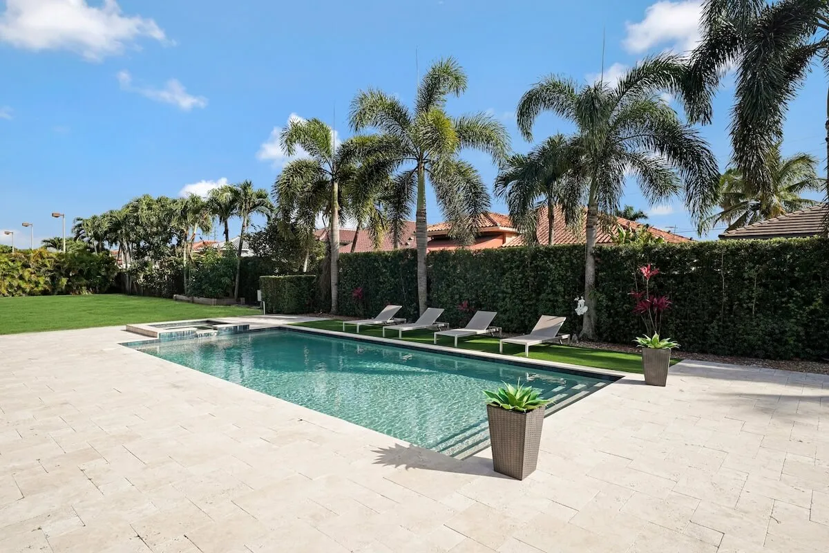 A rectangular swimming pool with crystal-clear water is surrounded by a row of four lounge chairs, perfect for sunbathing. Tall palm trees and neatly trimmed hedges frame the pool, creating a private and tropical atmosphere. The manicured green lawn offers additional space for games or gatherings.