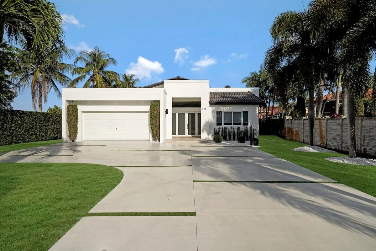 A striking view of the home's modern white facade, framed by tall palm trees and a large driveway with subtle grass strips. The inviting glass front door and well-maintained landscaping set a welcoming scene for arriving guests and create a strong first impression for any event.
